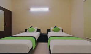 Une chambre d’hôtel avec deux lits simples, chacun recouvert d’une couvre-lit verte et blanche et doté d’un oreiller. Il y a une table de chevet avec un téléphone et une prise électrique. La chambre a un mur de couleur claire et une porte située sur le côté gauche.