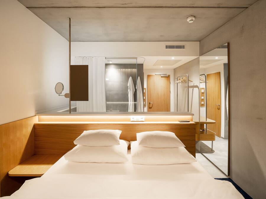 A modern hotel room with a large mirror, a wooden bench, and a bed with white linens.