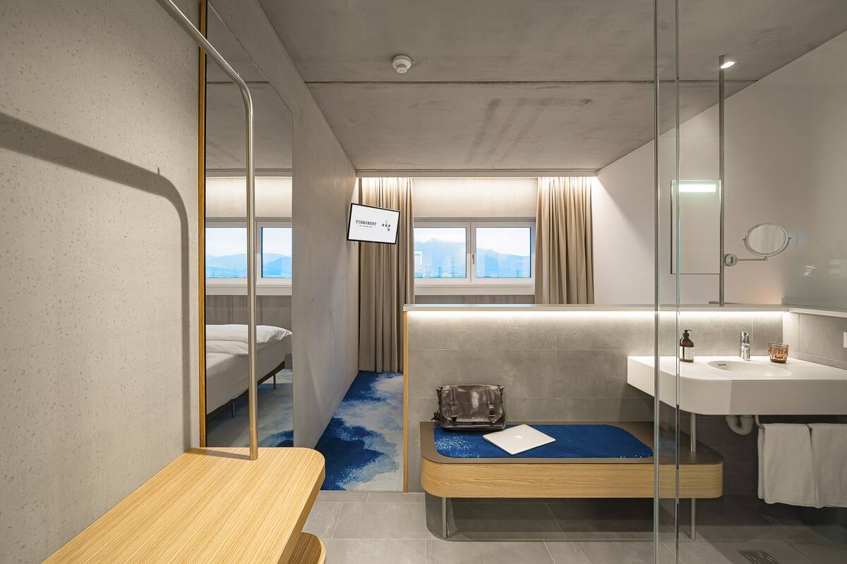 Habitación de hotel moderna con cama, un pequeño escritorio, un baño con lavamanos y espejo, y vistas a las montañas a través de la ventana.