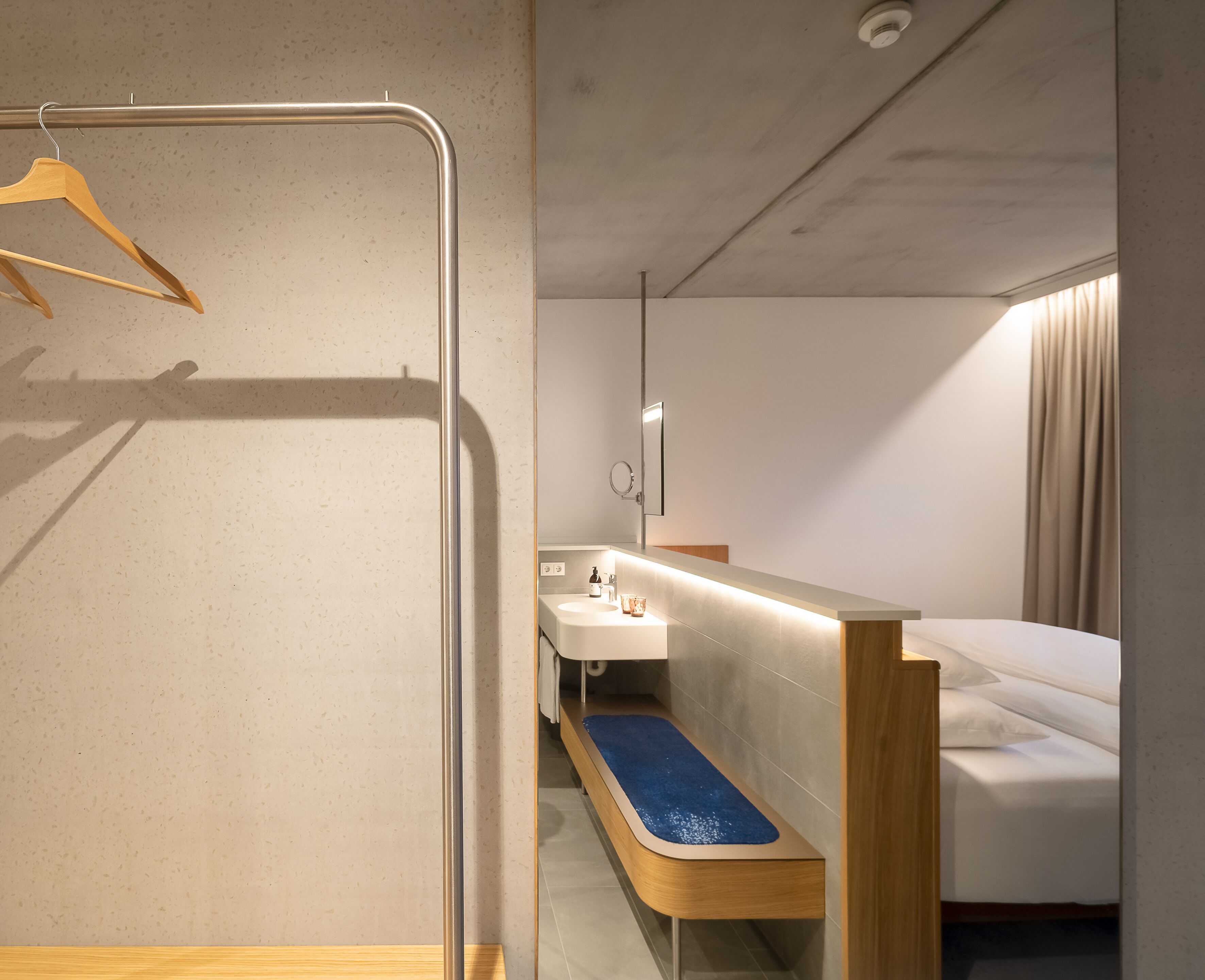 A modern hotel room with a wooden bench, a small sink, and a bed with a blue mat.