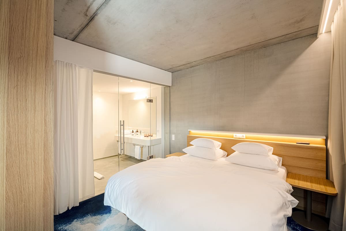 Una habitación de hotel moderna con una cama grande, una mesita de noche de madera, una lámpara de noche integrada y un baño visible a través de una puerta abierta.
