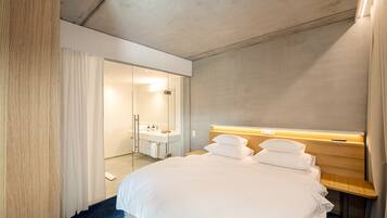 A modern hotel room with a large bed, a wooden bedside table, a built-in bedside lamp, and a bathroom visible through an open door.