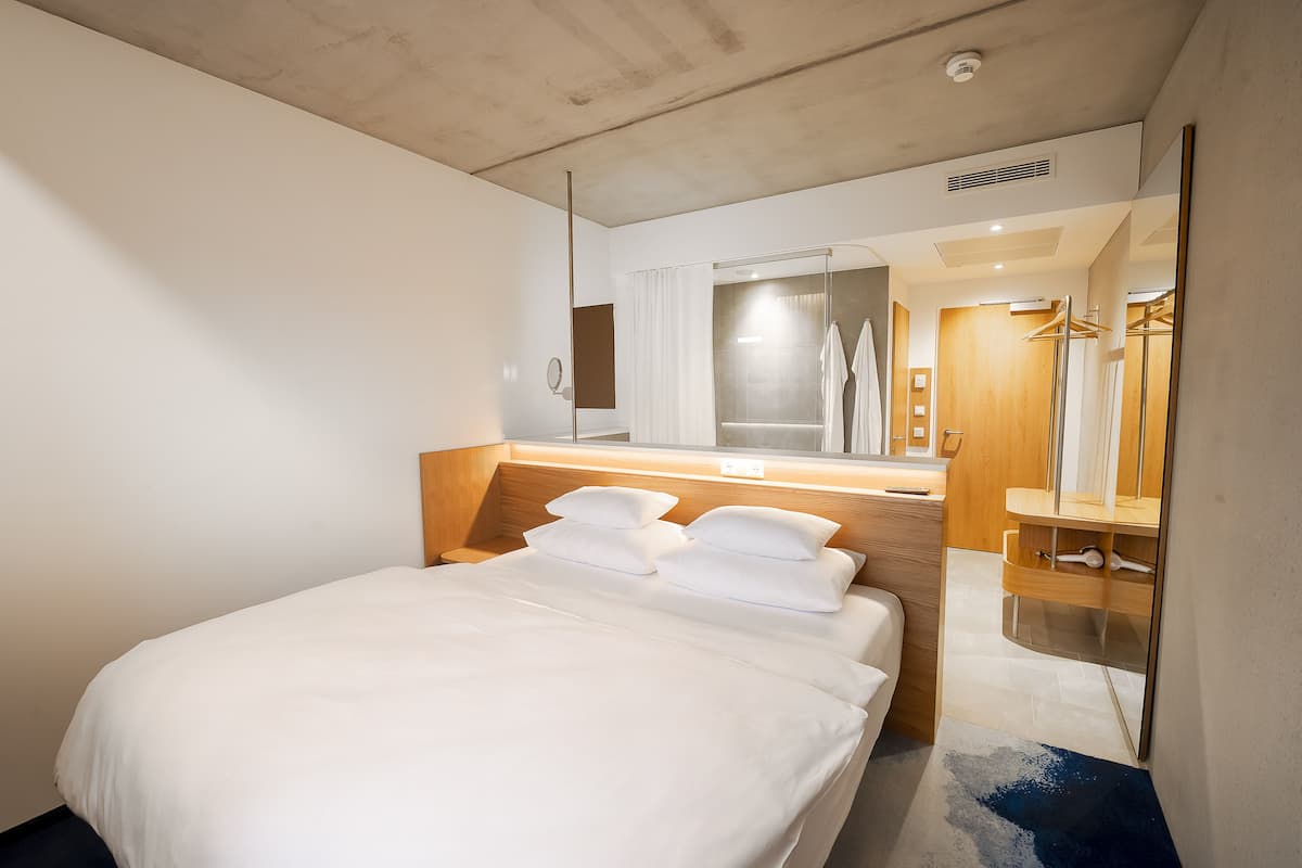 Una habitación de hotel moderna con una cama grande, un cabecero de madera, un espejo integrado, un tocador con espejo y una zona de ducha.