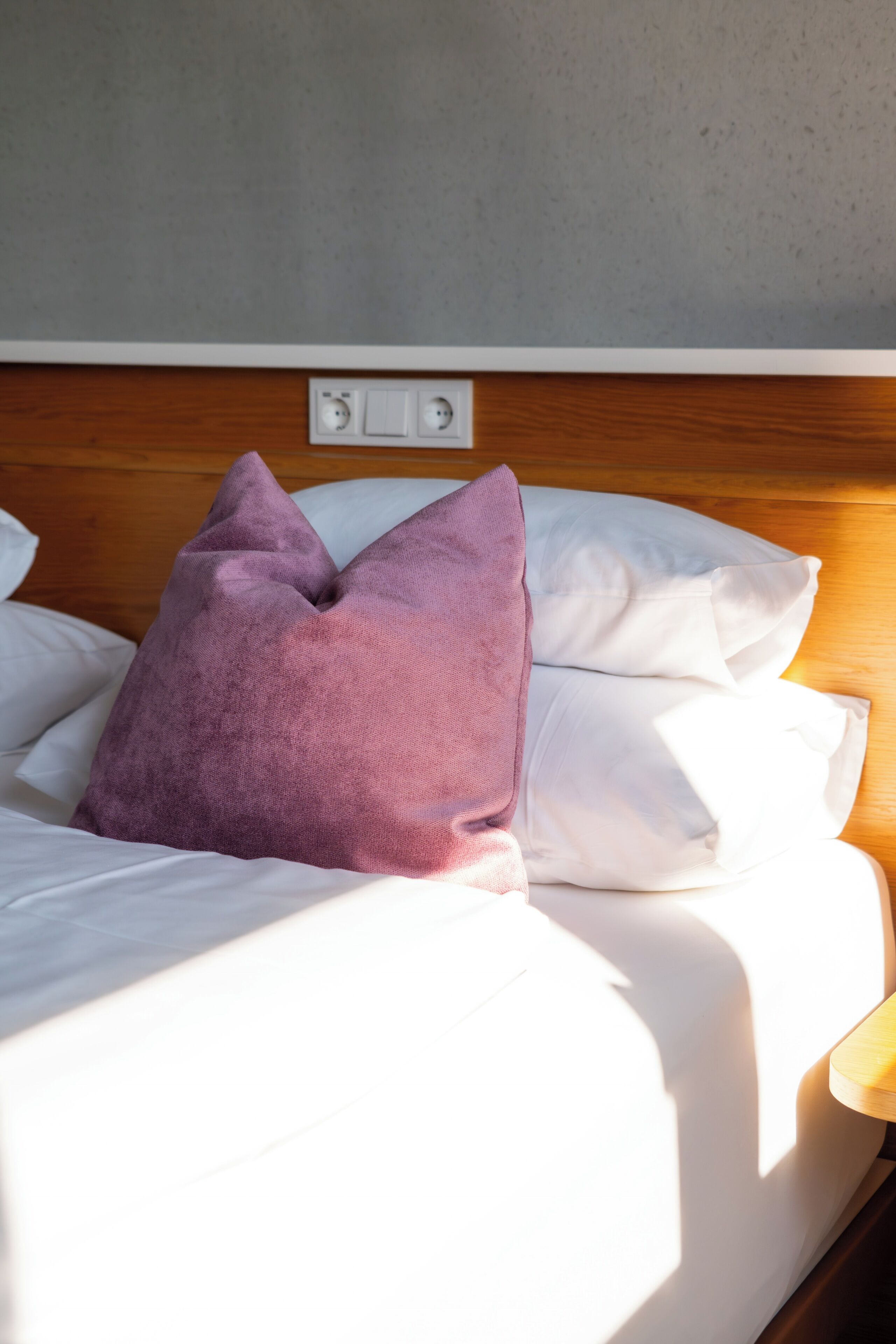 Uma cama com lençóis brancos, um travesseiro roxo e uma cabeceira de madeira com tomada.