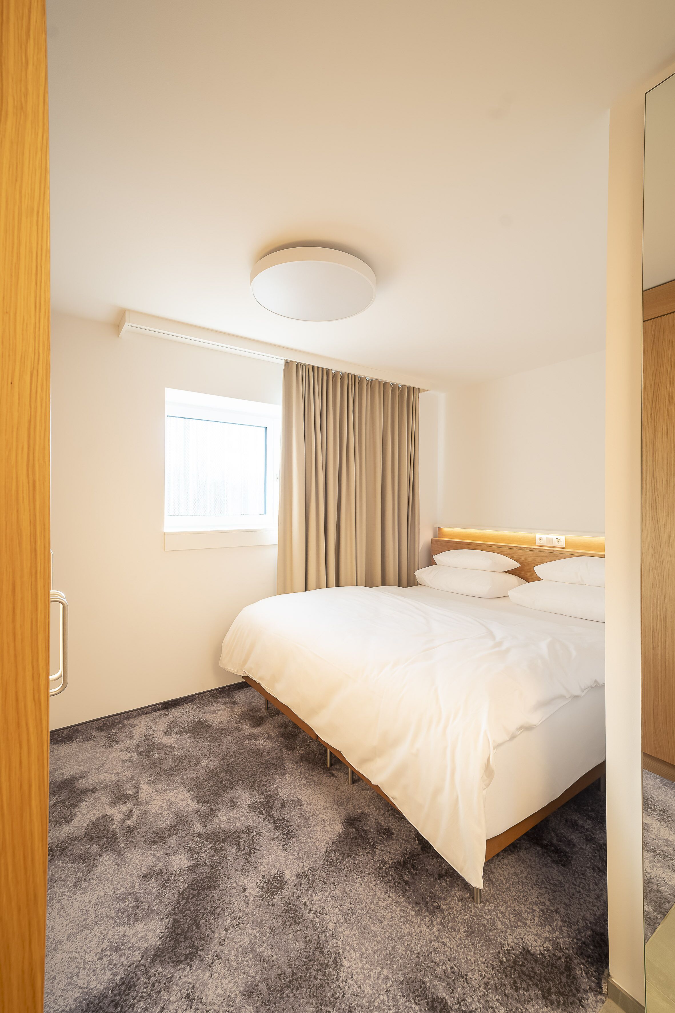 Habitación de hotel con una cama grande, una ventana con cortinas, una lámpara en el techo y piso alfombrado.