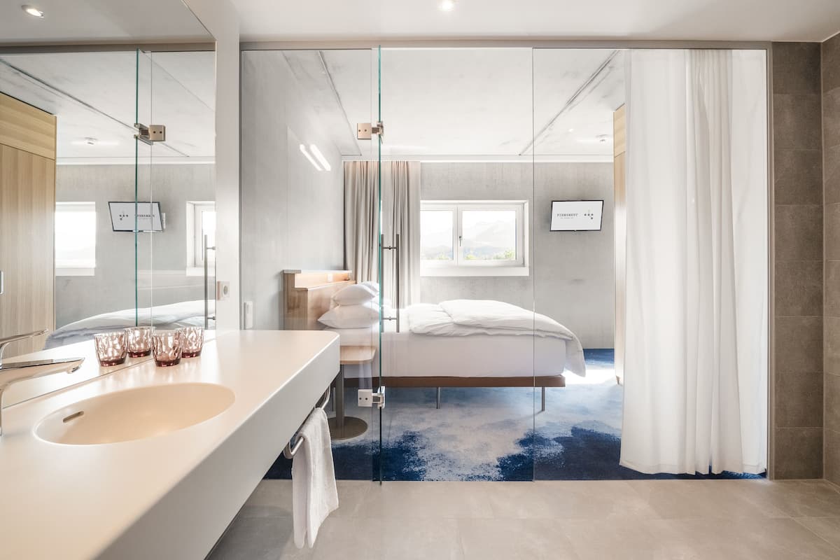 Una habitación de hotel moderna con un espejo grande, una cama blanca, un box de ducha de vidrio y vistas al exterior.