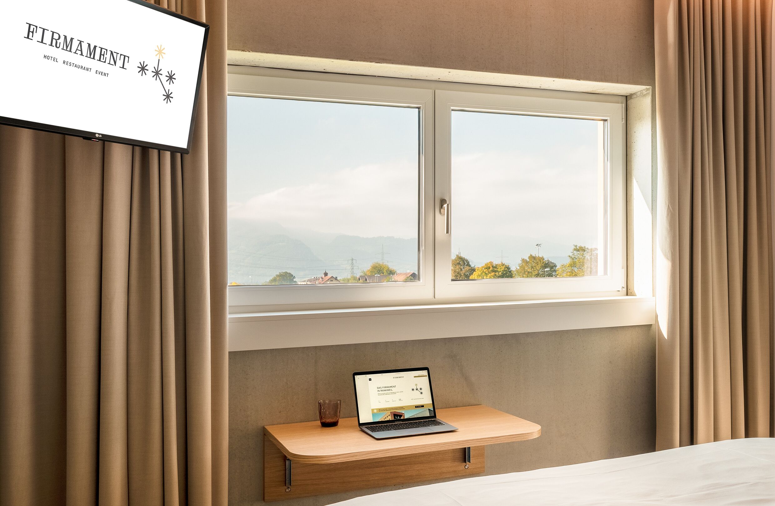 A hotel room with a bed, a desk with a laptop, a window with a view of mountains, and curtains.