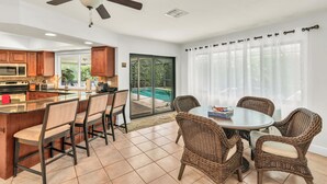 A kitchen with a central island, dining area with wicker chairs, and a view of a swimming pool through glass doors. - Short Walk to Maple Beach Access I 3/2 w/ Heated Pool (Marco Island)