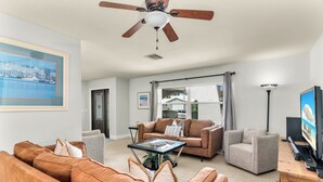 A living room with a brown leather sofa, a grey armchair, a glass coffee table, and a ceiling fan. - Short Walk to Maple Beach Access I 3/2 w/ Heated Pool (Marco Island)
