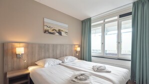 A hotel room with a large bed, two bedside tables, a headboard, a window with curtains, and a painting on the wall.