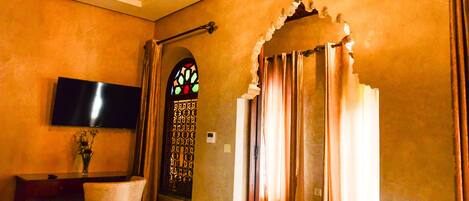 A room with a flat-screen TV mounted on the wall, a desk with a chair, a decorative stained glass window, and a door with curtains.