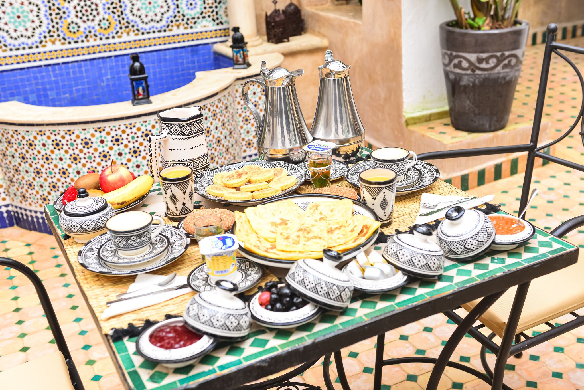 A table set with traditional Moroccan tea and food, including pastries and fruits, in a tiled outdoor area.