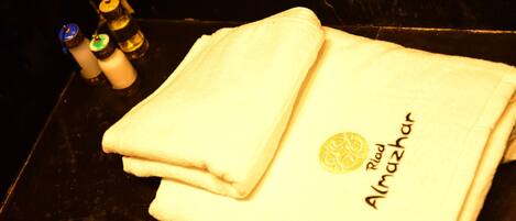 A folded white towel with "Riad Almazhar" embroidered on it, placed on a black surface next to three bottles.