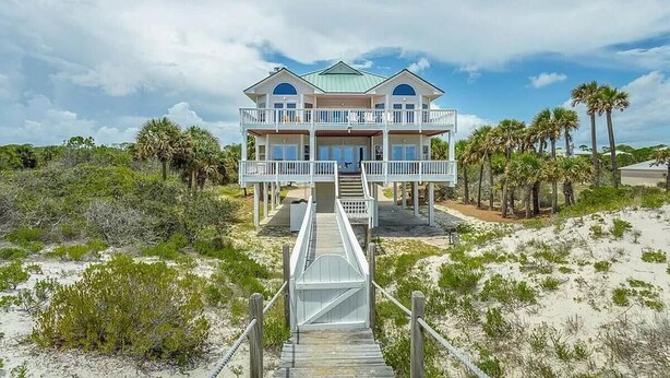 A beachfront house with a balcony, a wooden deck, and a view of the beach and palm trees.