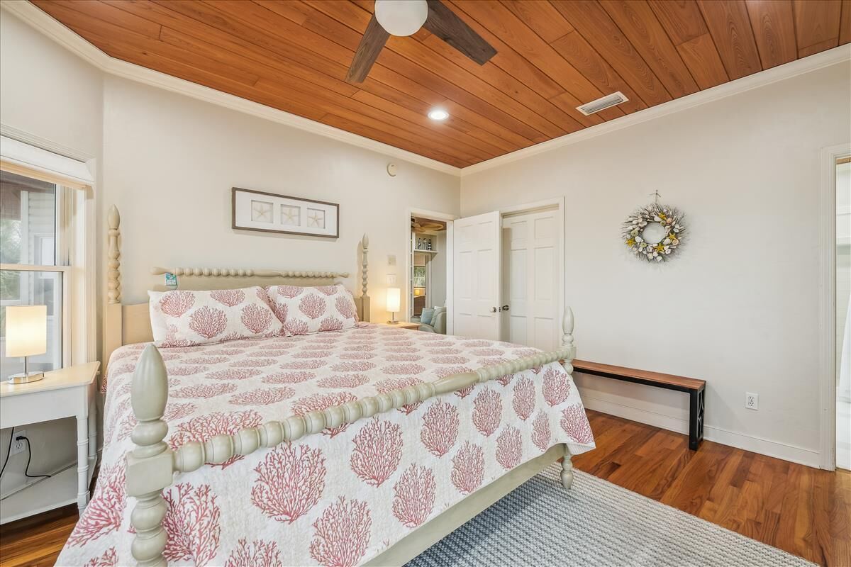 A bedroom with a wooden ceiling, a bed with a patterned bedspread, a bedside table with a lamp, a bench, and a door with a wreath.