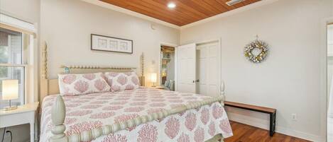 A bedroom with a wooden ceiling, a bed with a patterned bedspread, a bedside table with a lamp, a bench, and a door with a wreath.