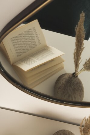 Un livre ouvert sur une surface blanche, avec un miroir qui reflète le livre et un vase contenant des plantes séchées.