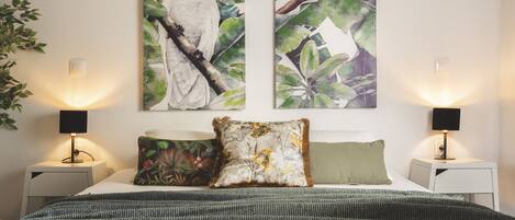 Une chambre à coucher comprenant un lit, deux tables de chevet, deux lampes fixées au mur et deux tableaux représentant des oiseaux.