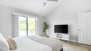 Een moderne slaapkamer met een groot bed, een televisie op een witte kast, en een glazen schuifdeur die leidt naar een buitenruimte.
