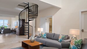 Un vaste espace de vie agrémenté d’un escalier moderne, d’un canapé d’angle gris, d’une table basse en bois et d’une petite table d’appoint ronde en bois sur laquelle repose un verre de vin.