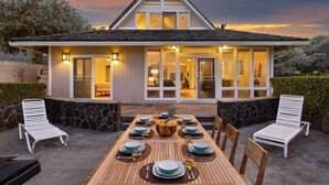 Un coin repas avec une table en bois dressée pour six personnes, entourée de chaises blanches, avec un mur en pierre et une maison en arrière-plan.