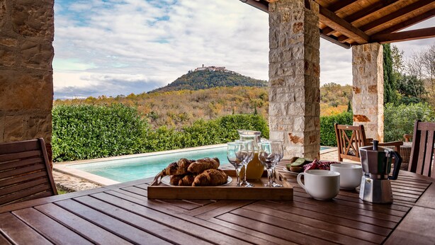 Une table en bois, dressée avec des aliments pour le petit-déjeuner et du matériel à café, donnant sur une piscine et une colline sur laquelle se trouve un bâtiment.