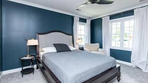 Un dormitorio con cama, mesita de noche, silla, ventilador de techo y ventana con cortinas.