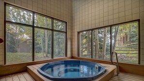 A hot tub in a corner with large windows, wooden flooring, and a view of trees and a building. - Spacious Mountain Retreat with Private Balcony Offering Slope and River Views (Keystone)