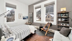 A bedroom with a bed, a chair, a shelf with books and a lamp, and a window with a view of a snowy landscape.