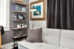 Een woonkamer met een grijze bank, een gekleurd sierkussen, een boekenplank met boeken en decoratieve objecten, een ingelijst schilderij en een vloerlamp.