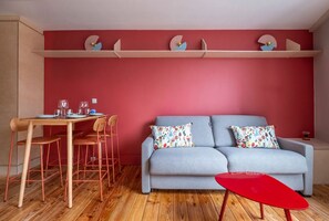 A living room with a grey sofa, two patterned pillows, a red coffee table, a wooden dining table with chairs, and a red wall with decorative elements.