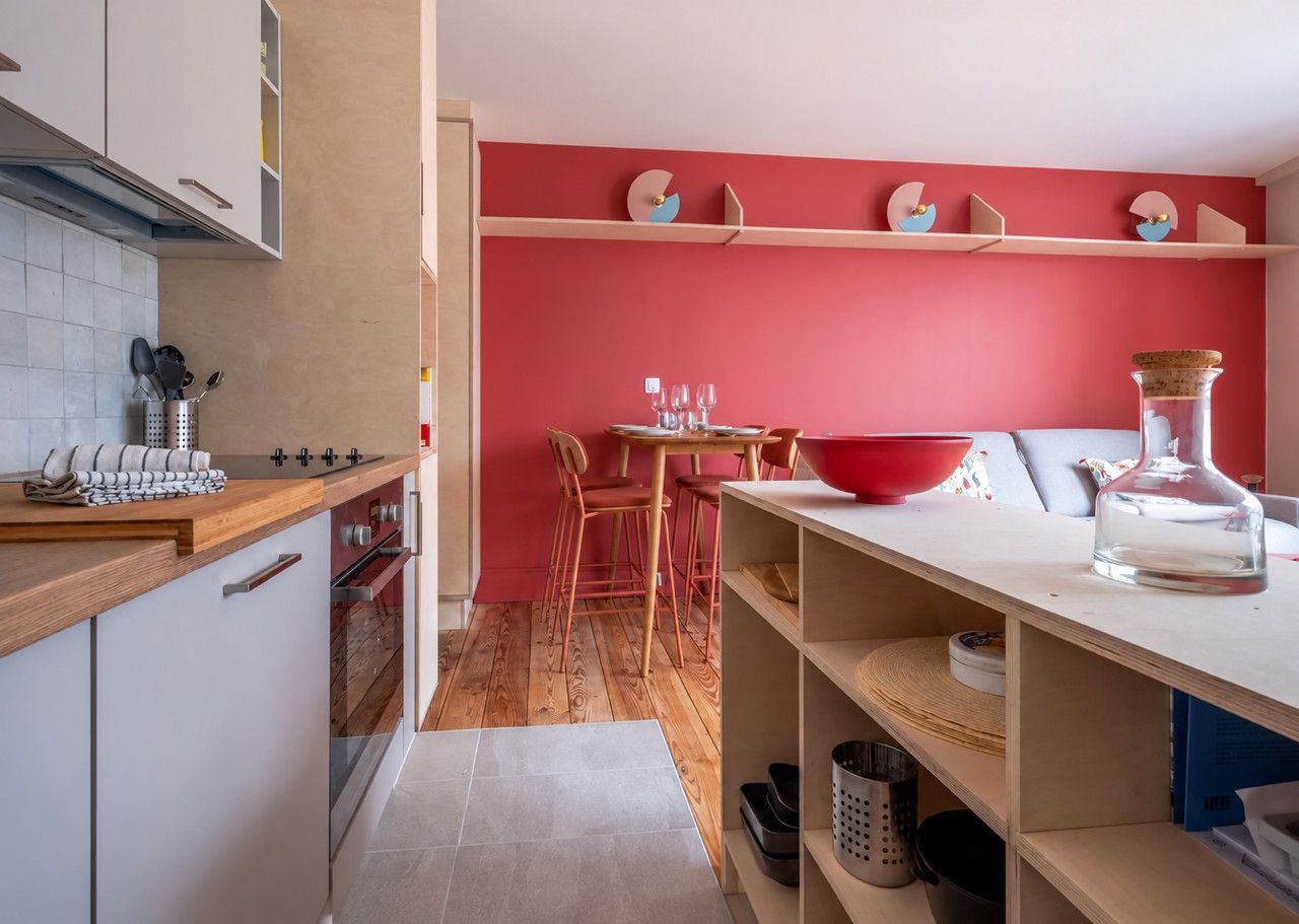 A modern kitchen with a red accent wall, wooden cabinets, and a dining area with red chairs.