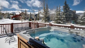 Un jacuzzi con cubierta de madera y sillas metálicas, rodeado de árboles nevados y montañas al fondo.
