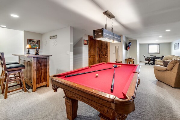 A pool table with a red felt surface, wooden chairs, and a bar area with a lamp.