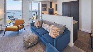 A modern living room with a blue sofa, a yellow armchair, and a patterned ottoman. There is a balcony with a view of the sea. - Host & Stay | The Waves (Whitby)