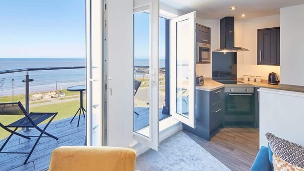 A modern kitchen with an open layout, a balcony with a view of the sea, and a seating area with a sofa and armchair. - Host & Stay | The Waves (Whitby)