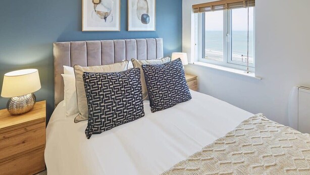 A bedroom with a large bed, two patterned pillows, a bedside lamp, a wooden nightstand, and a window with a view of the beach.