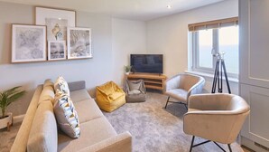 A modern living room with a sofa, two armchairs, a coffee table, and a TV. There are framed artworks on the wall and a tripod in the corner.