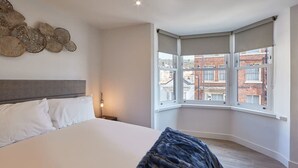 A bedroom with a large bed, a headboard, a wall art piece, a window with a view of buildings, and a corner window with a view. - Host & Stay | Othala @ The Merchants Quarters (Scarborough)