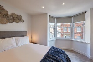 A bedroom with a large bed, a headboard, a wall art piece, a window with a view of buildings, and a corner window with a view. - Host & Stay | Othala @ The Merchants Quarters (Scarborough)