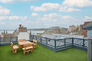 A rooftop terrace with a wooden table and chairs, surrounded by a decorative metal fence, overlooking a cityscape with buildings and a body of water.