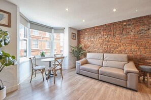 A modern living room with a brick wall, a beige sofa, wooden flooring, and a dining area with chairs and a table.