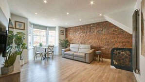 A spacious living area with a brick wall, a sofa, a dining table with chairs, and potted plants. - Host & Stay | Othala @ The Merchants Quarters (Scarborough)