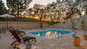 Un espace extérieur avec une piscine dont le sol est en pierre, agrémenté de chaises longues et d’une table, le tout entouré d’arbres et d’un mur en pierre.