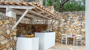 Un espace cuisine extérieur doté de murs en pierre, d’une pergola et d’une table avec des chaises.