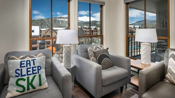 Una sala de estar moderna con un conjunto de sofá, dos lámparas y vista a un paisaje montañoso nevado a través de amplias ventanas.