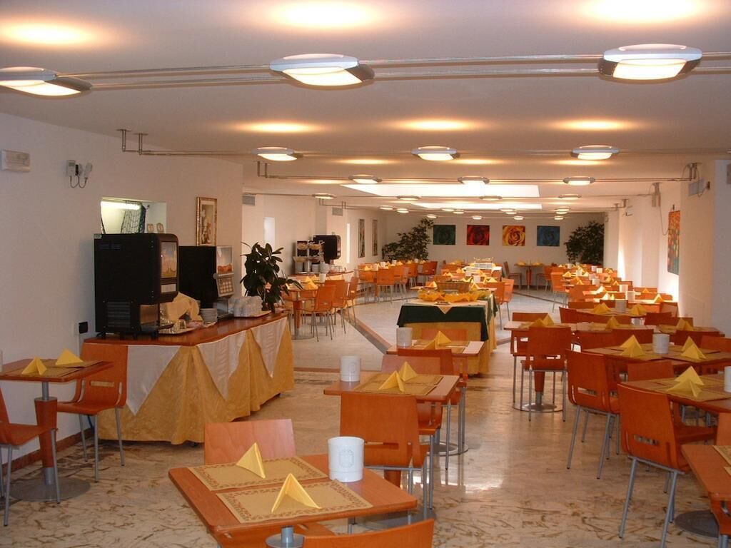 A spacious dining area with multiple tables and chairs, a counter with a cash register, and a large plant in the center.