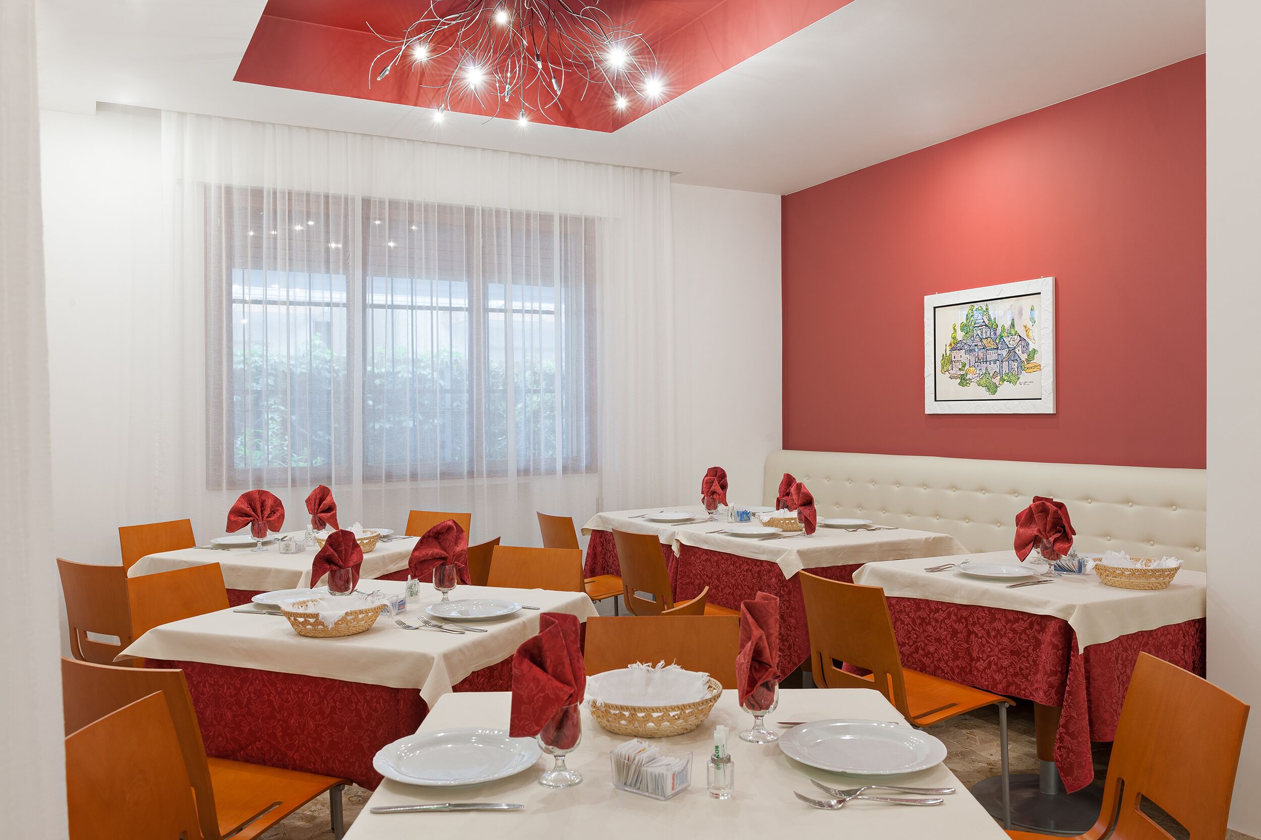 Un espace repas avec des tables dressées pour quatre personnes, des nappes blanches, des serviettes rouges et un mur d’accent rouge.