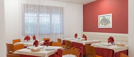Un espace repas avec des tables dressées pour quatre personnes, des nappes blanches, des serviettes rouges et un mur d’accent rouge.