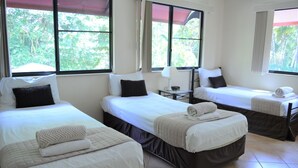 Habitació d'hotel amb tres llits individuals, cadascun amb un coixí i una tovallola arrotllada. Hi ha finestres amb cortines i una petita taula de nit amb una làmpada entre els llits.
